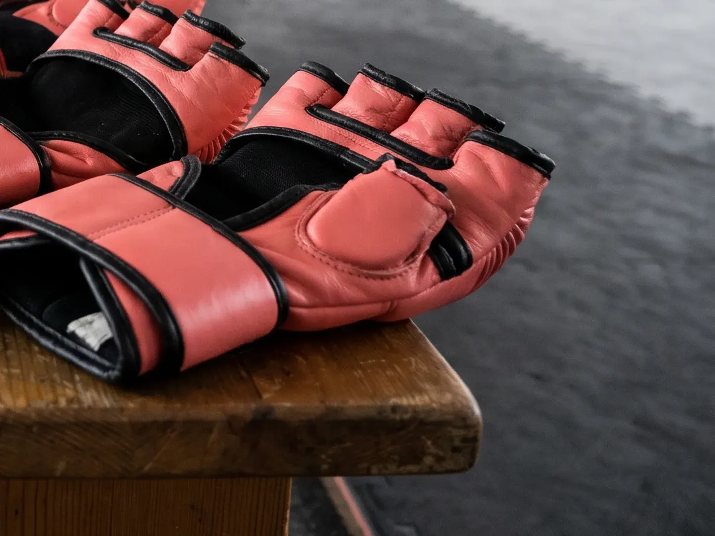 Coral and black fingerless MMA bag gloves laid on a worn wooden gym bench, showing the open finger openings, thick knuckle padding and long wrist straps.