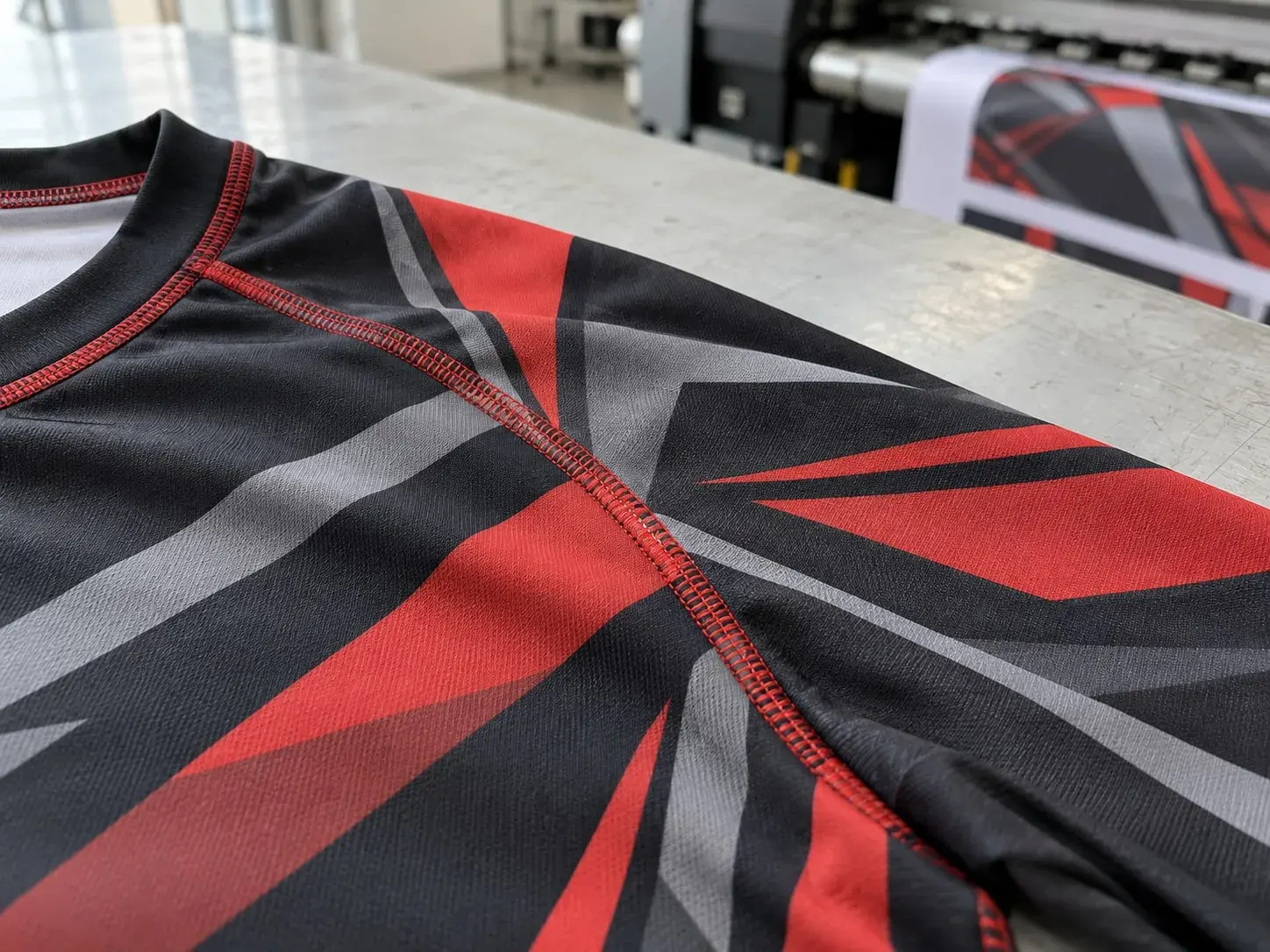 A black rashguard printed edge to edge with red and grey geometric panels, the sublimated print running across a seam, a printer feeding out transfer paper behind.