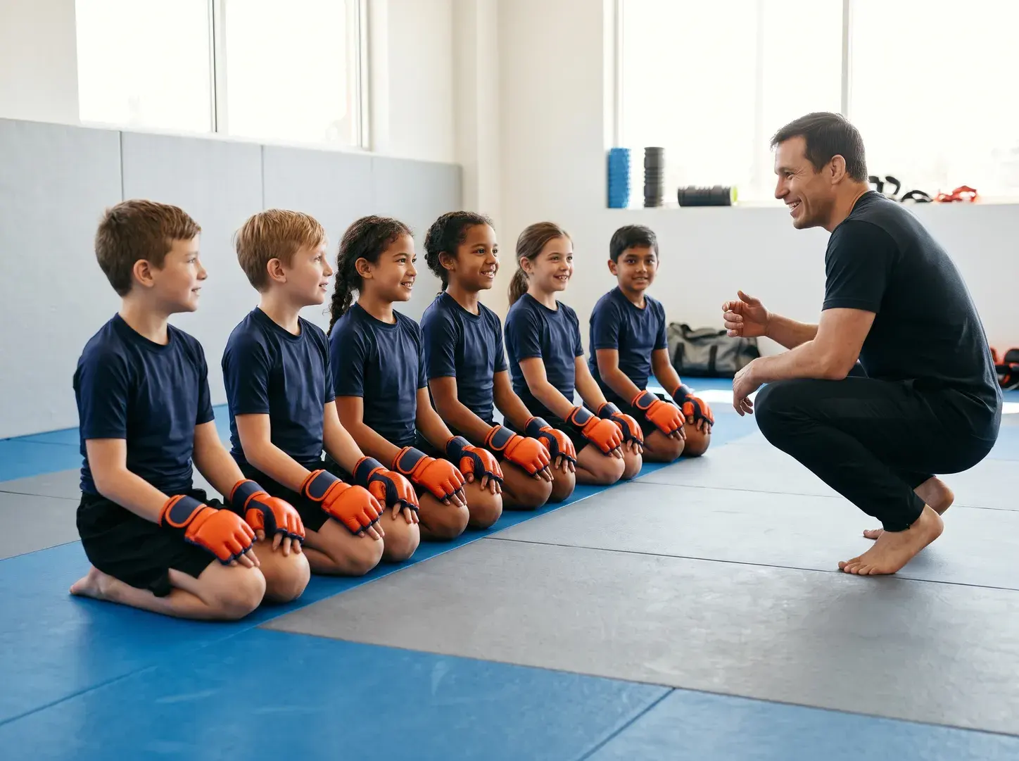 Six children kneeling in a line on blue and grey mats in matching bright orange and navy kids' MMA gloves and navy rash guards, smiling at their coach, who crouches in front of them.