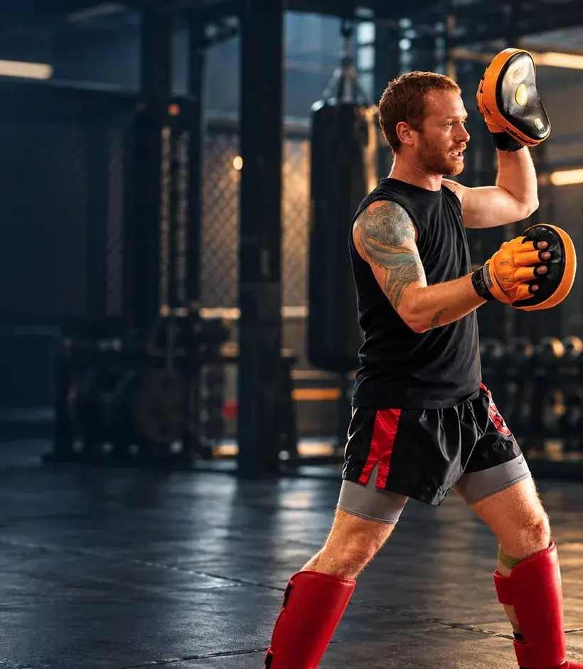 A coach in a black vest and red shin guards holding up focus mitts in the gym.
