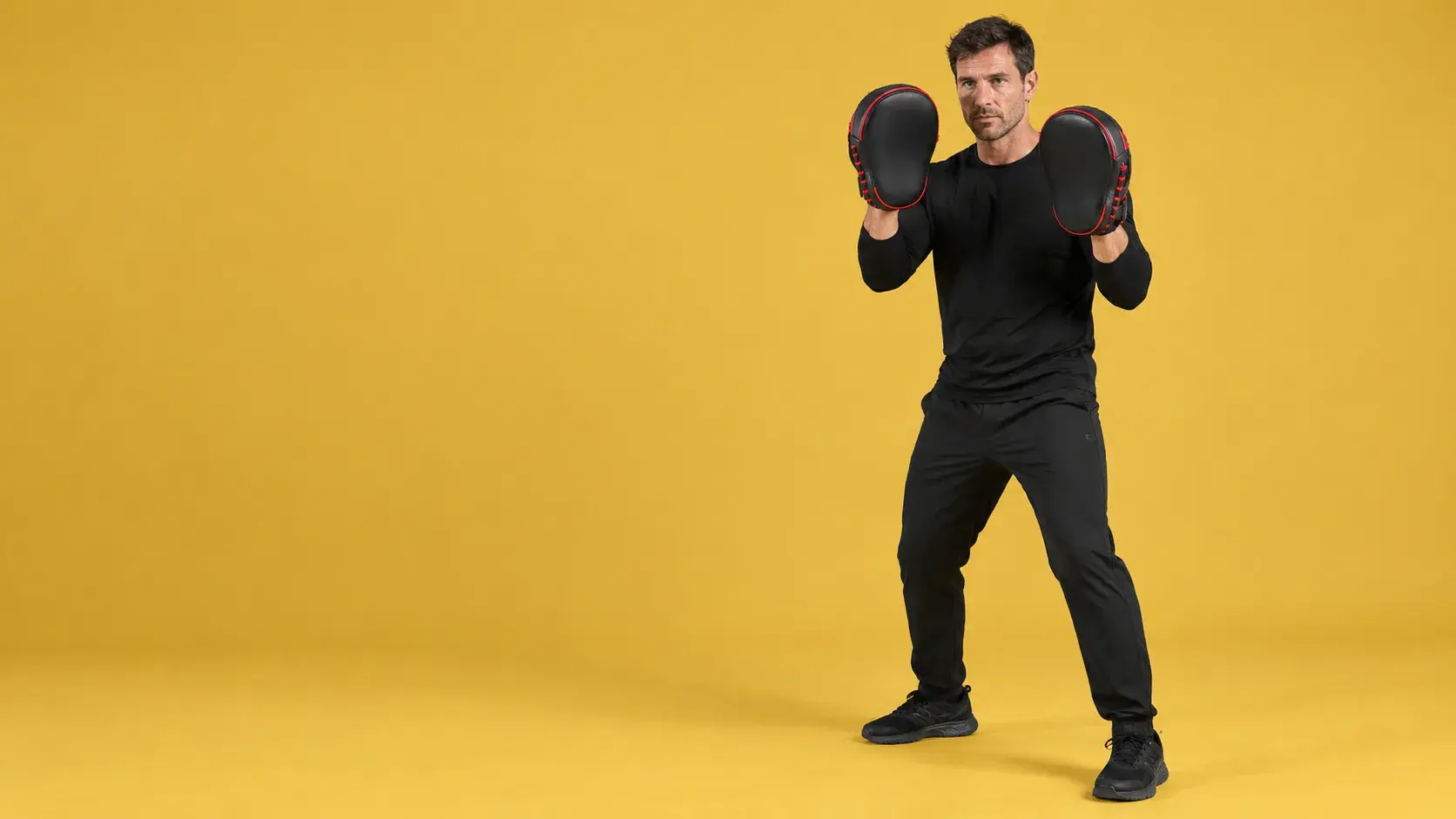 A coach on a mustard yellow backdrop holding up a pair of black and red curved focus mitts as targets.