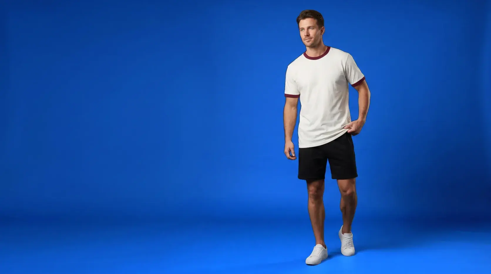 A man on a royal blue backdrop in an off-white T-shirt with a burgundy ringer collar and sleeve cuffs, black shorts and white trainers.