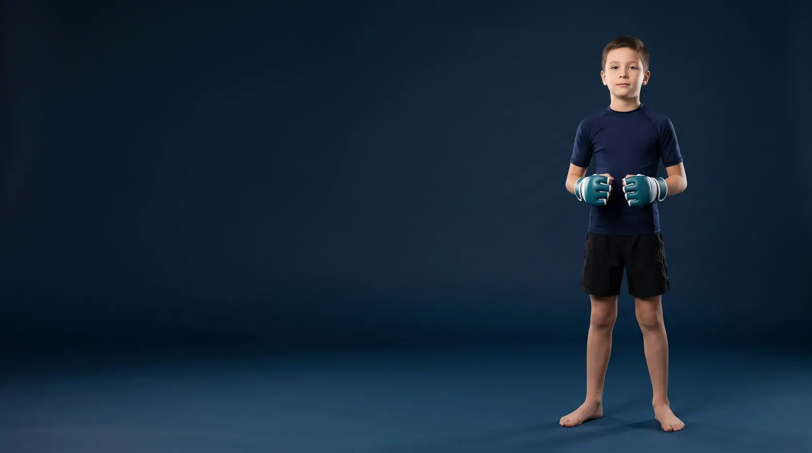 A child standing on a midnight navy backdrop in small teal and white open-palm kids' MMA gloves, a navy rash guard and black shorts, barefoot.