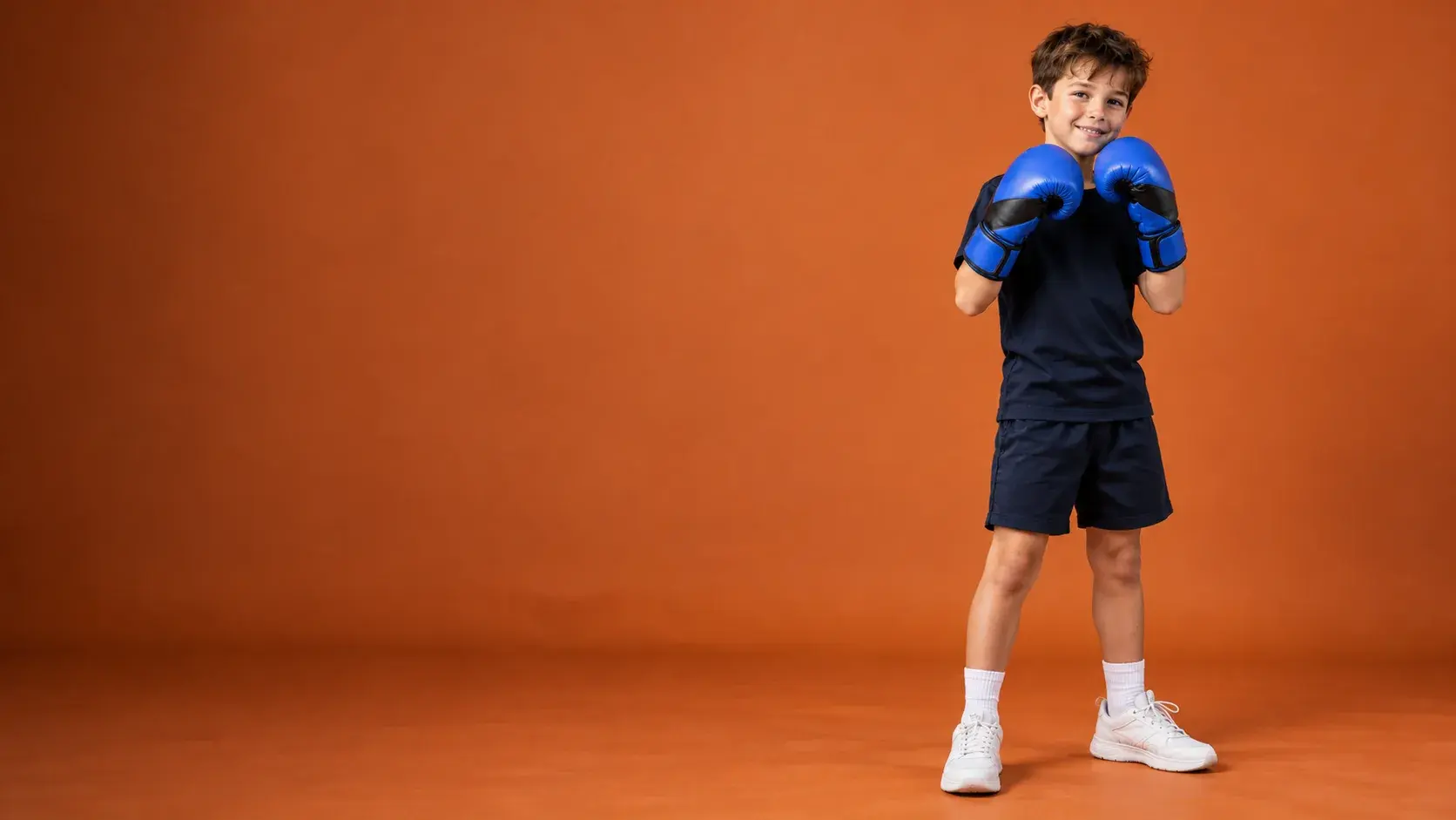 A junior boxer in a guard on a burnt orange backdrop, wearing royal blue and black kids' boxing gloves, a navy T-shirt, navy shorts and white trainers.
