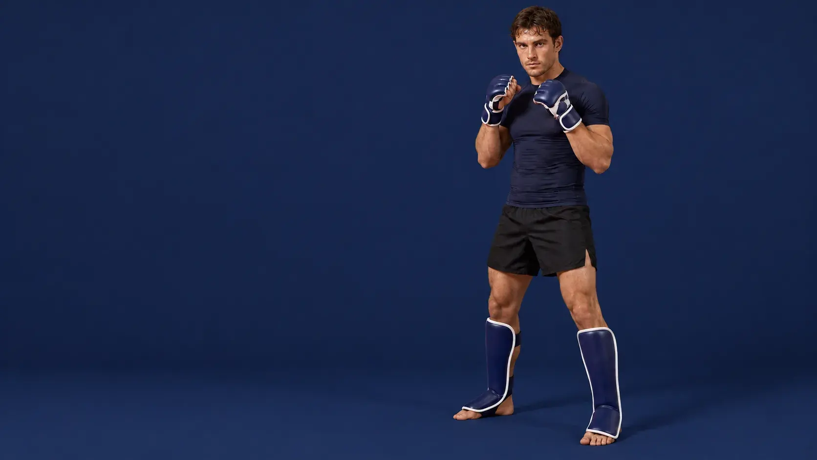 An MMA fighter in a fighting stance on a midnight navy backdrop, wearing navy and white open-palm sparring gloves, navy shin-and-instep guards, a navy rash guard and black fight shorts.