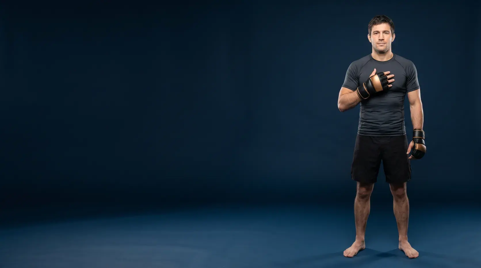 An MMA fighter standing on a midnight navy backdrop with one gloved hand raised to the chest, in black and bronze open-finger MMA bag gloves, a charcoal rash guard and black fight shorts, barefoot.