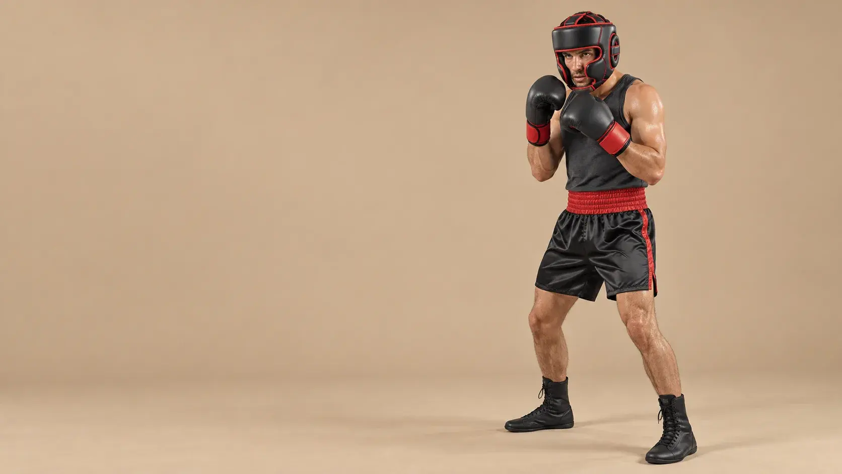 A boxer in a guard on a warm sand backdrop, wearing a black cheek-protector head guard with red piping, black gloves with red cuffs, a charcoal vest, black satin shorts and black boots.