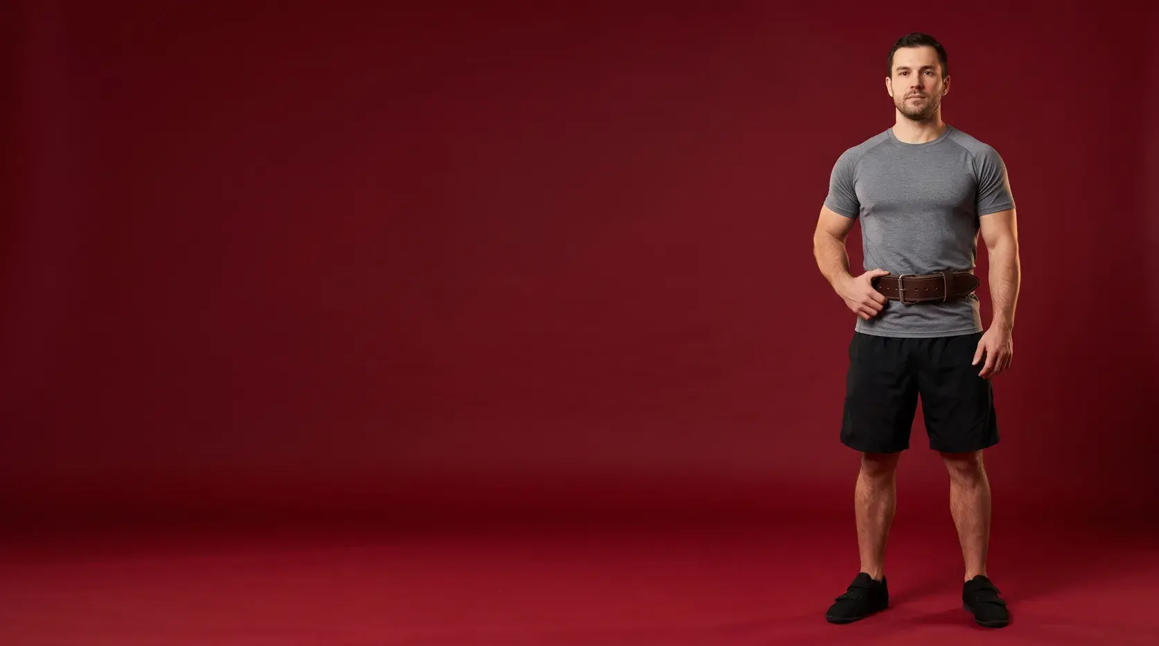 A strength athlete standing on an oxblood backdrop in a dark brown single-prong leather lifting belt, one hand resting at the hip, in a grey training top and black shorts.