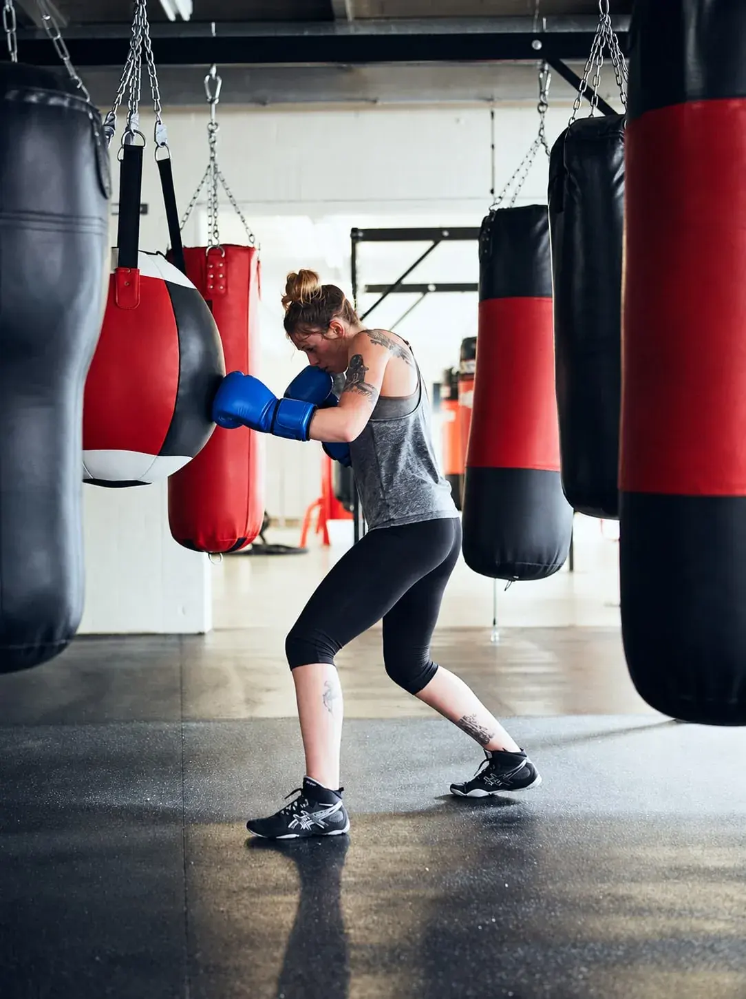 A boxer in blue gloves working a red and black teardrop bag in a gym hung with heavy bags.