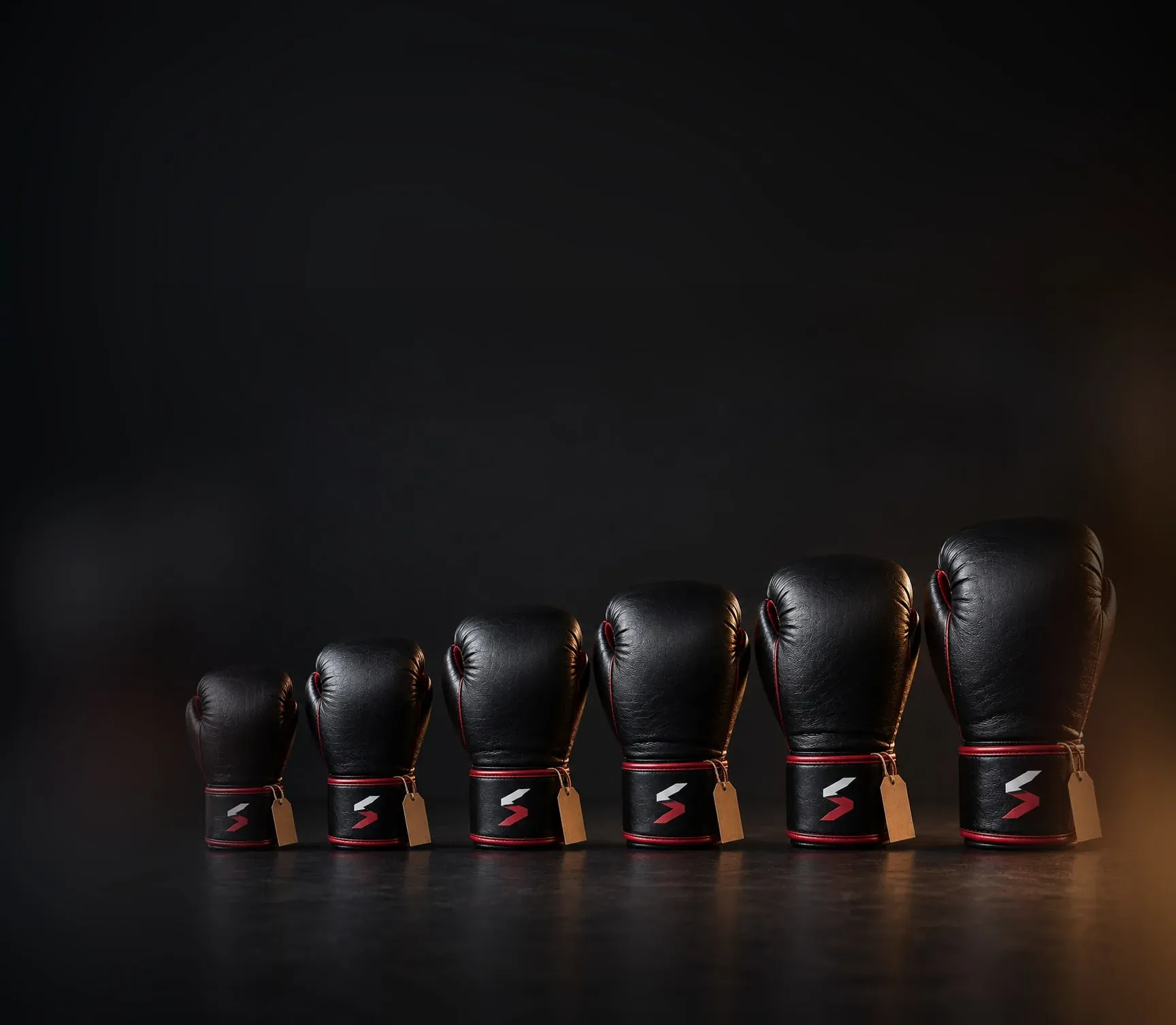 The same black and red boxing glove with the Sparwright logo on the cuff in a row of weights, from the smallest kids’ glove to the largest adult glove, each with a blank swing tag.