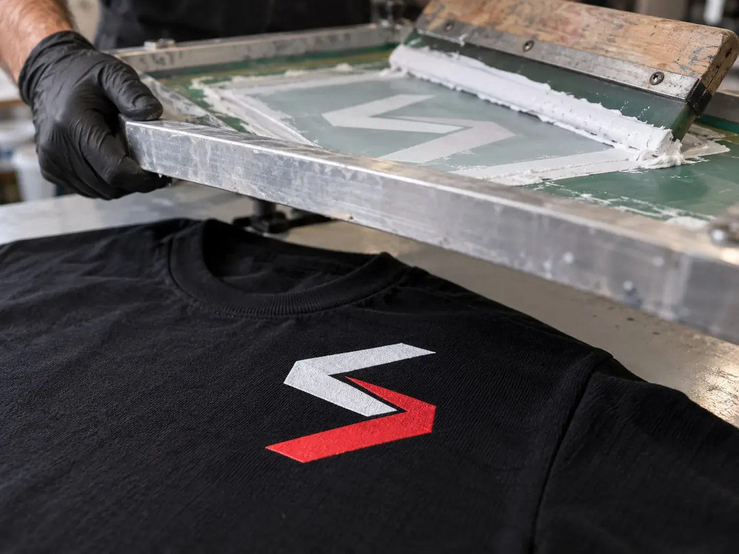 A gloved hand lifting a screen-printing frame off a black T-shirt, the Sparwright logo freshly printed on the chest in white and red, the squeegee resting in white ink on the screen.