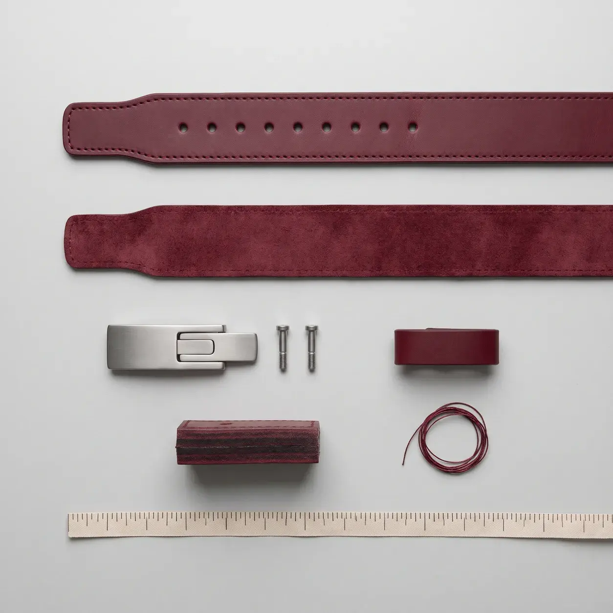 A burgundy leather lever belt laid out in its parts: the outer belt with its row of holes, the suede inner lining, the brushed steel lever buckle and its two screws, the keeper loop, a thickness sample and thread, with a tape measure.