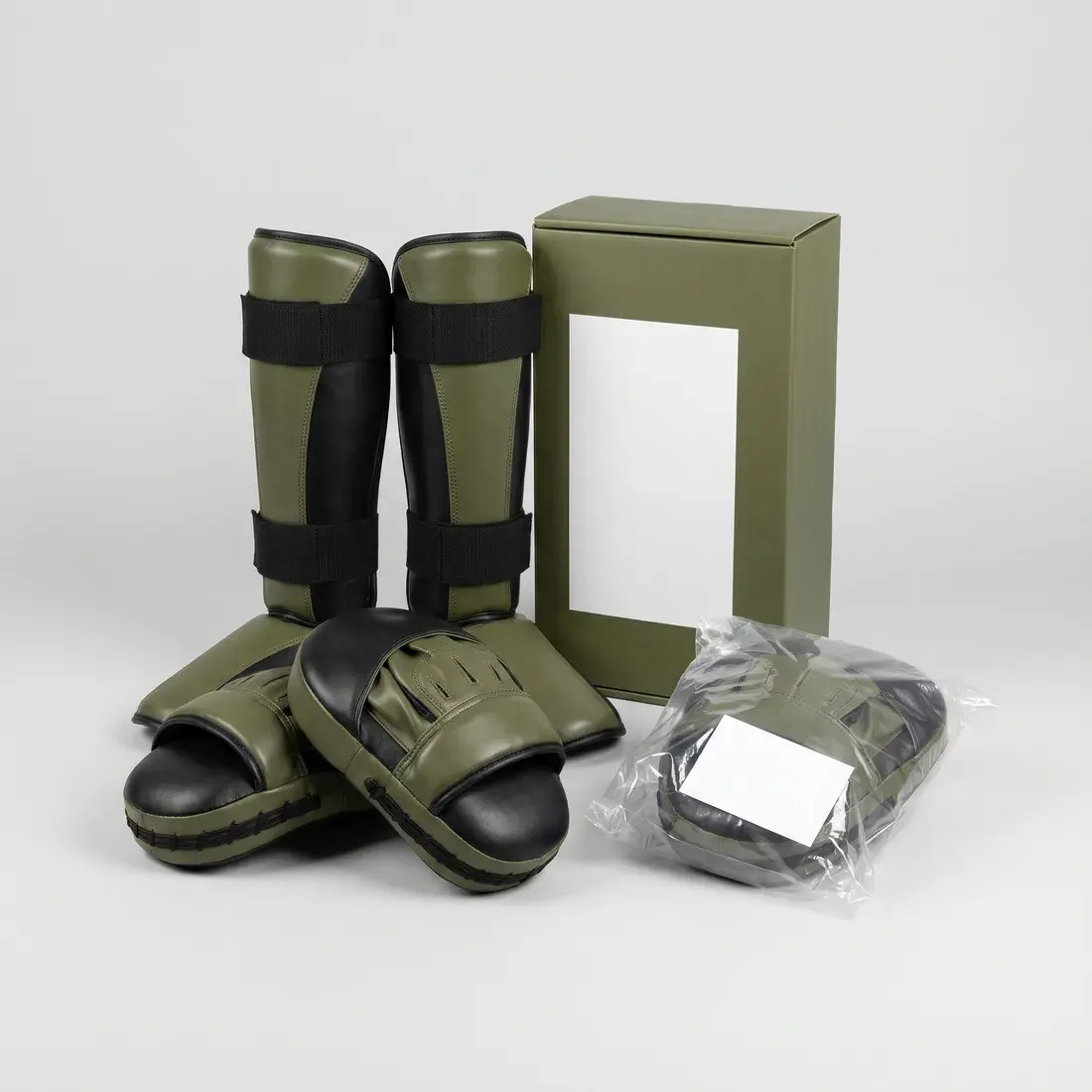 A pair of olive and black shin-and-instep guards and a pair of focus mitts, beside an olive retail box with a blank label panel and a pair of mitts in a labelled polybag.