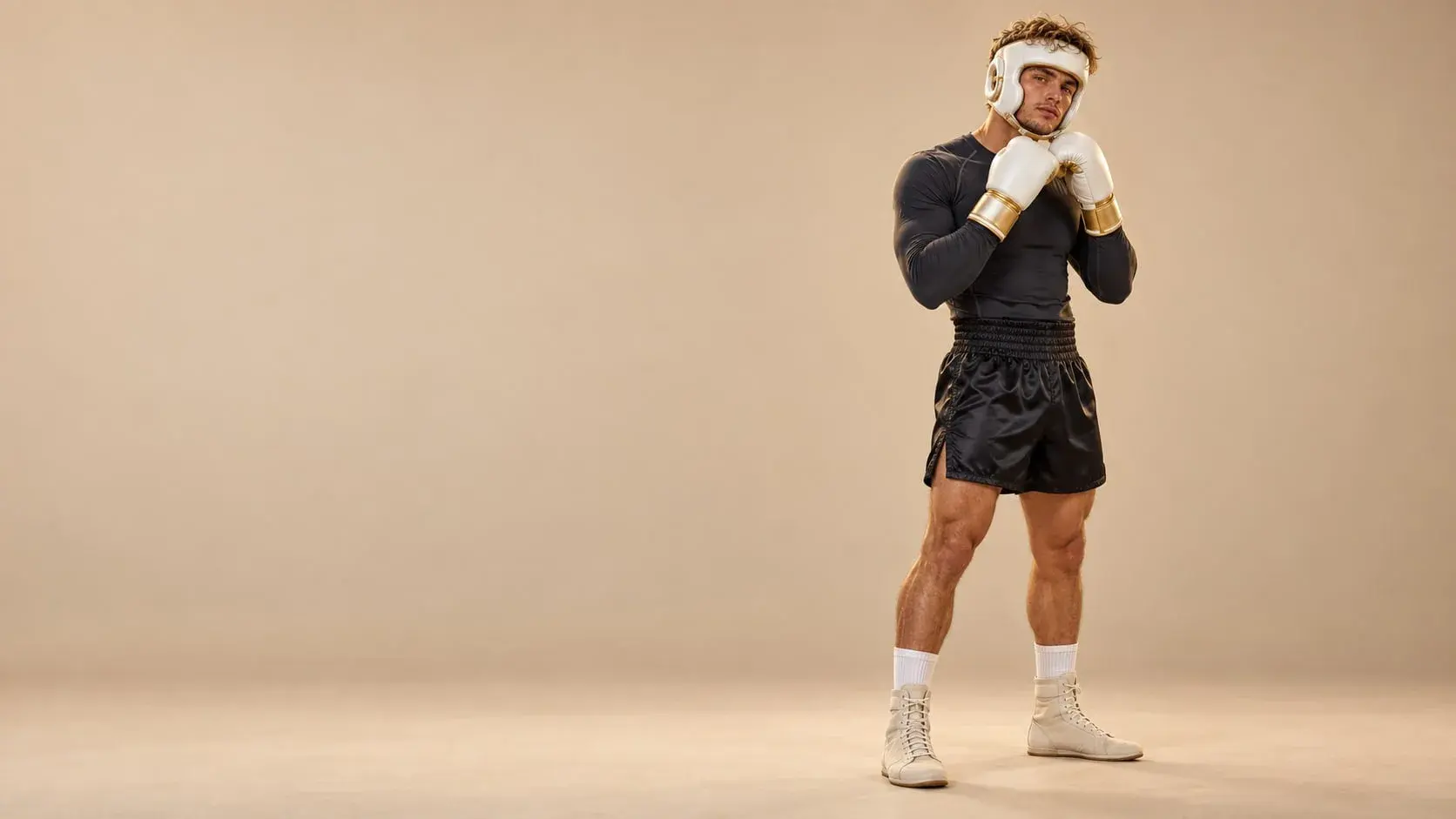 A boxer on a warm sand backdrop in a white open-face head guard with gold piping, white gloves with gold cuffs, a charcoal rash guard and black satin shorts.