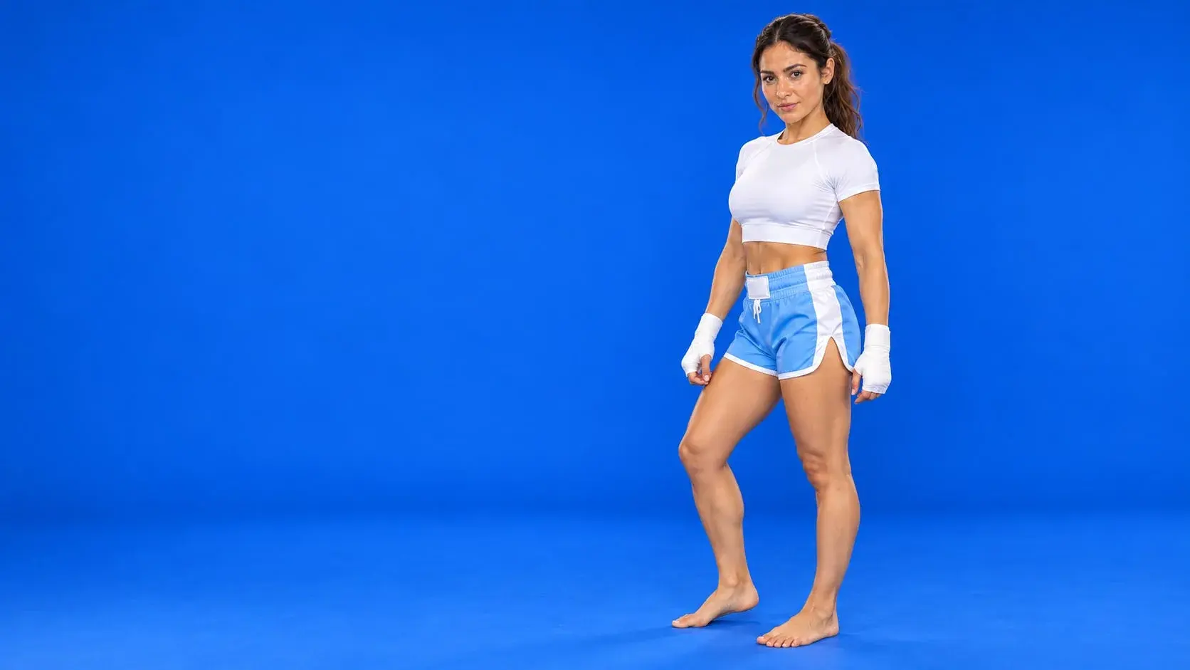 A fighter on a royal blue backdrop in sky blue fight shorts with white side panels and waistband, a white compression top and white hand wraps.