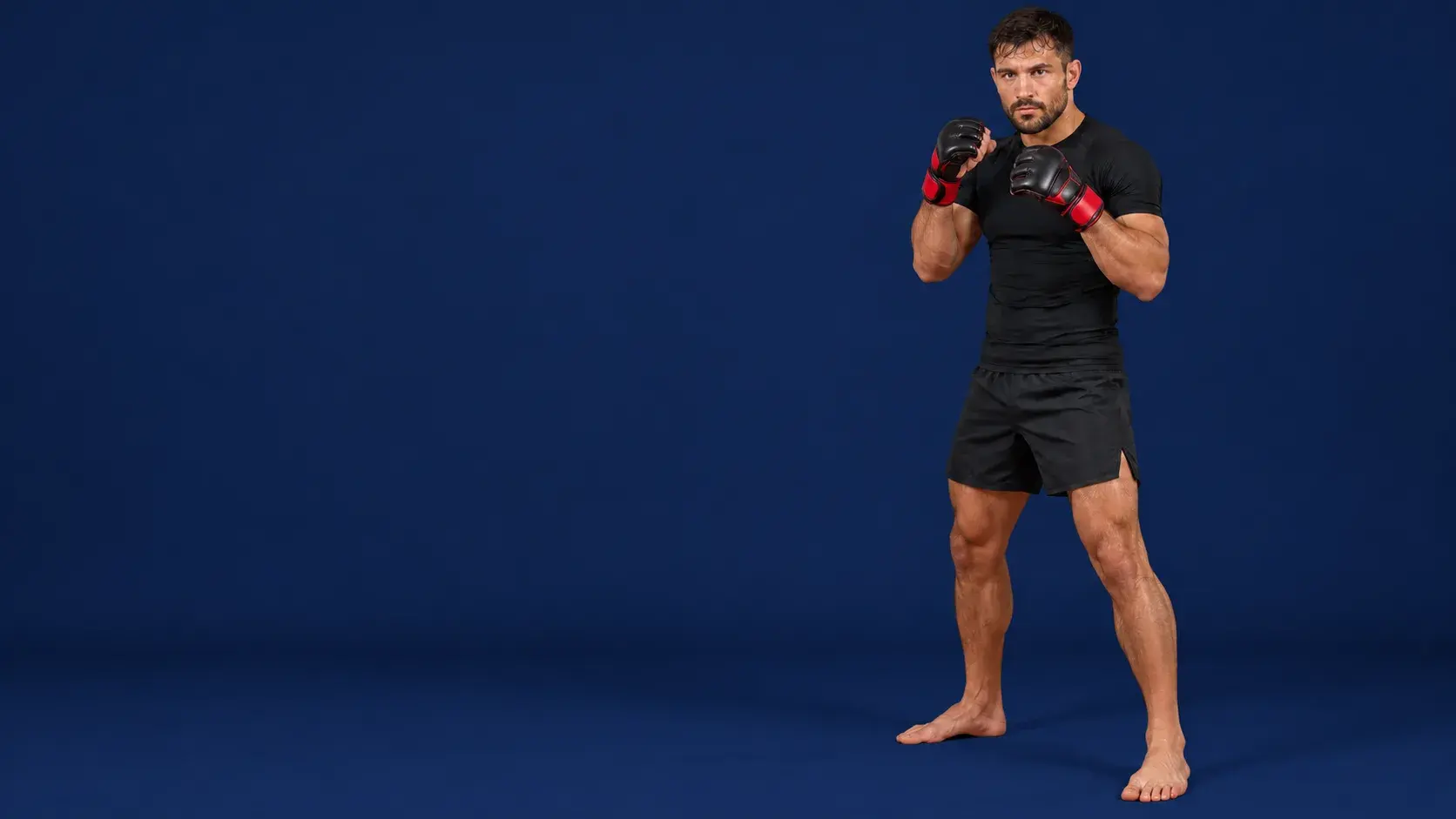 An MMA fighter in a fighting stance on a midnight navy backdrop, wearing black and red open-palm fight gloves, a black rash guard and black fight shorts, barefoot.
