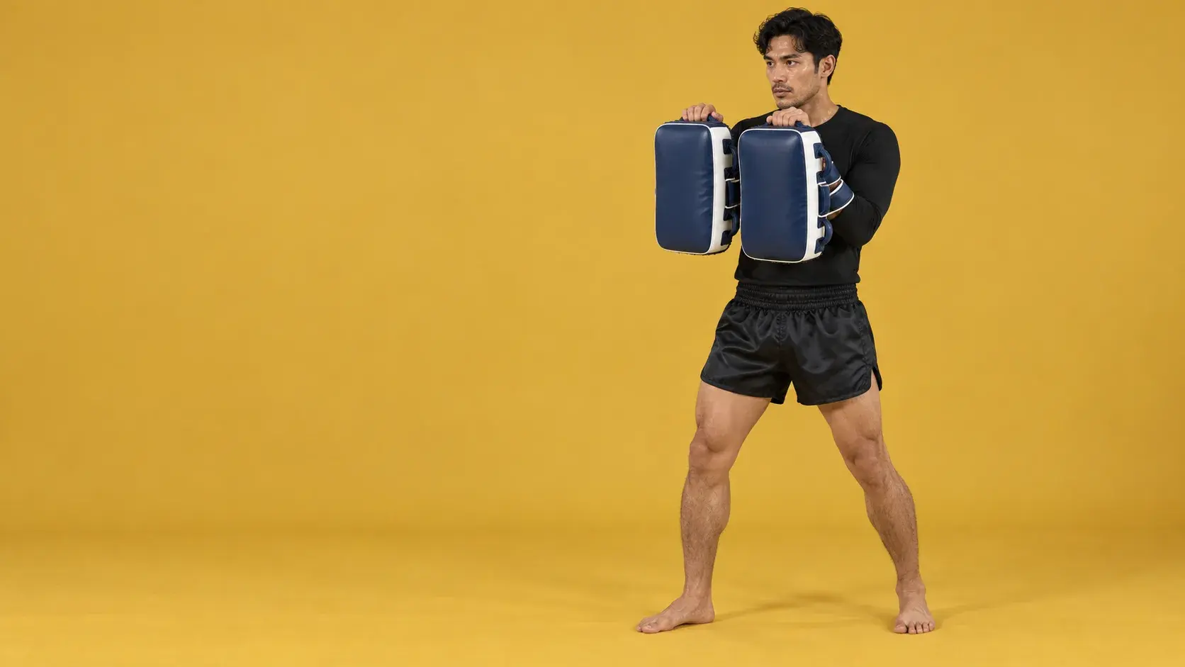 A pad holder on a mustard yellow backdrop holding a pair of navy and white Thai pads up in front of him.