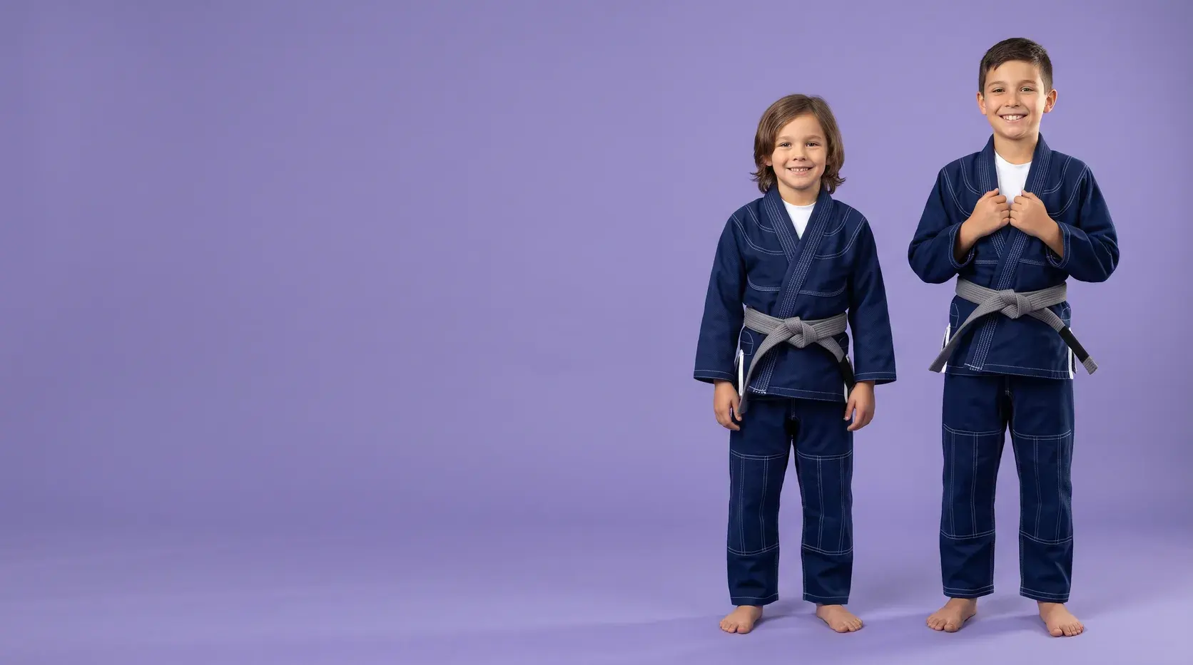 Two children on a violet backdrop in matching navy kids' BJJ gis with contrast stitching, tied with grey belts.