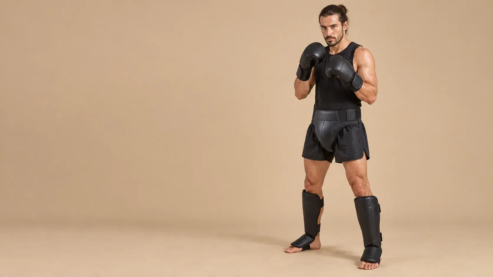 A kickboxer in a guard on a warm sand backdrop, wearing a black groin guard over black shorts, black gloves, a black vest and black shin-and-instep guards.