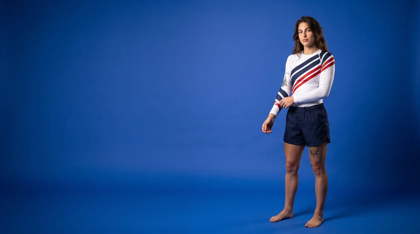 A grappler on a royal blue backdrop in a white long-sleeve rash guard with navy and red diagonal stripes and navy shorts, pulling down a cuff.