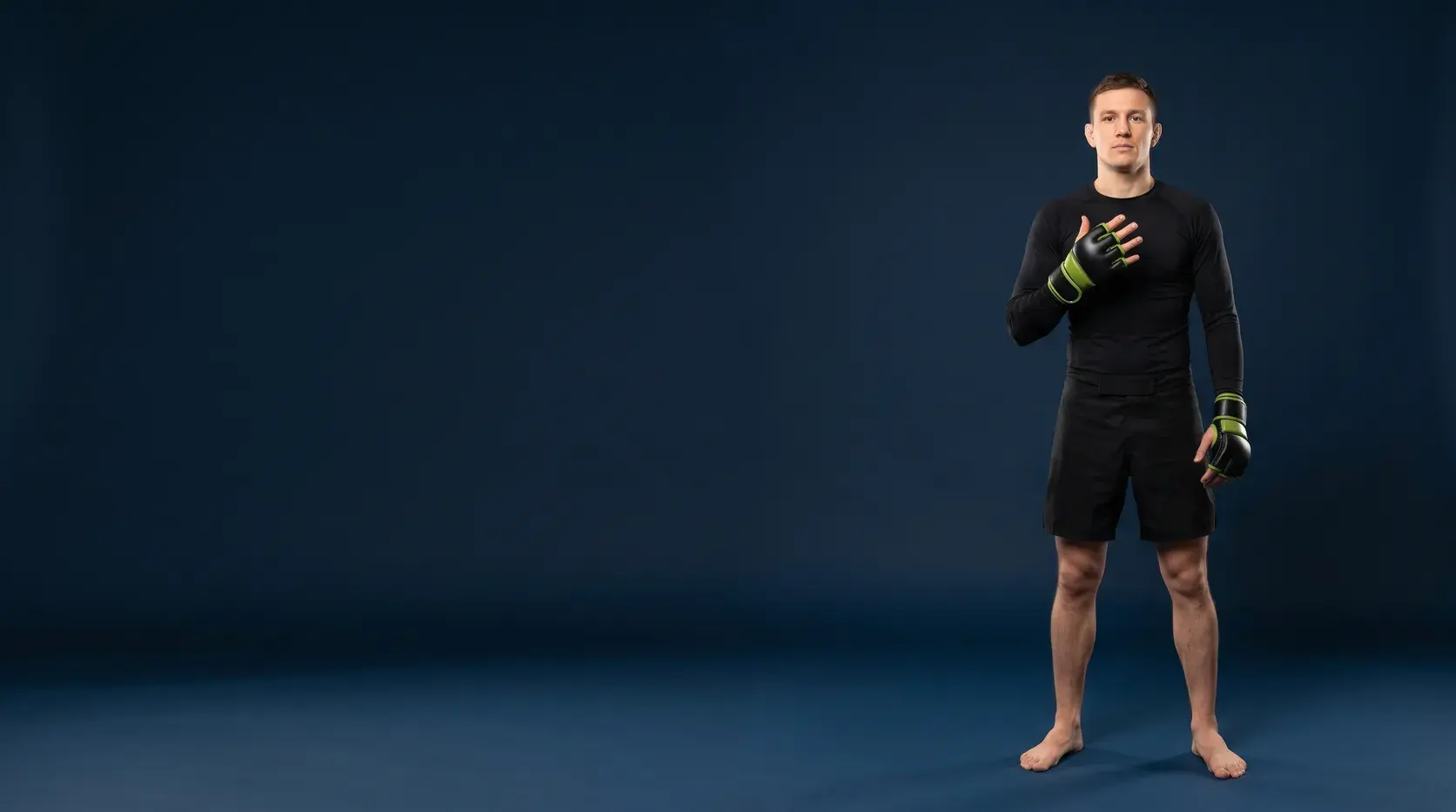 A grappler standing on a midnight navy backdrop with one open hand raised to the chest, in black and lime green grappling gloves with exposed fingers, a black long-sleeve rash guard and black fight shorts, barefoot.
