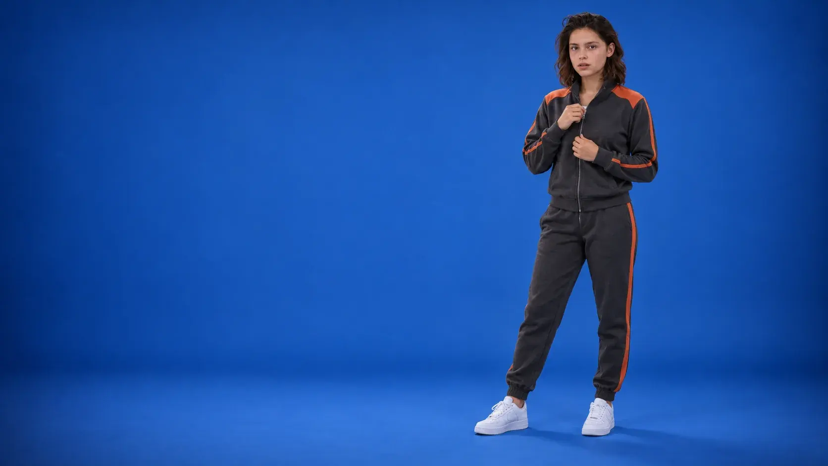 A young woman on a royal blue backdrop zipping up a charcoal tracksuit jacket with burnt orange shoulder panels and piping, with matching charcoal joggers with an orange side stripe and white trainers.