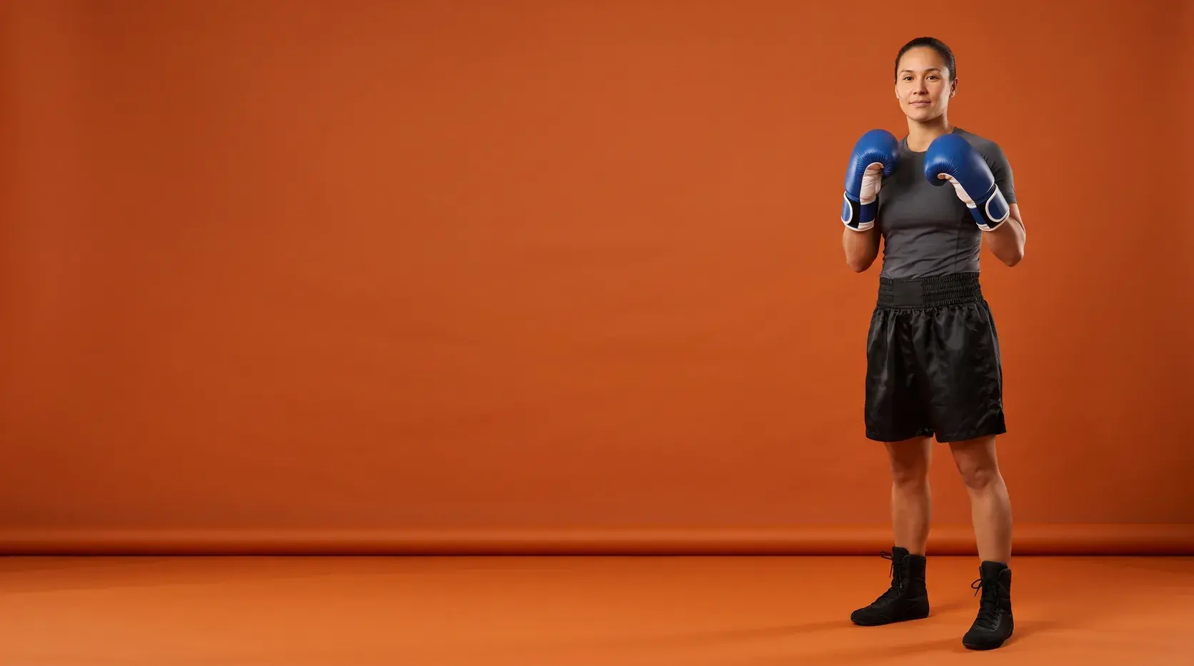 A boxer in a relaxed guard on a burnt orange backdrop, wearing royal blue and white hook-and-loop training gloves, a charcoal short-sleeve top, black satin shorts and black boots.
