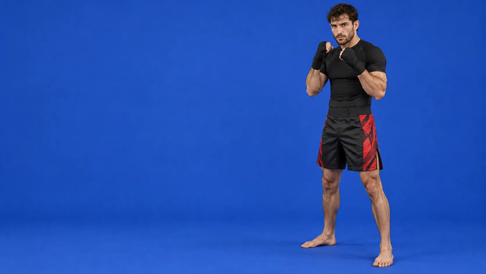 A fighter on a royal blue backdrop in black and red MMA fight shorts, a black rash guard and black hand wraps.