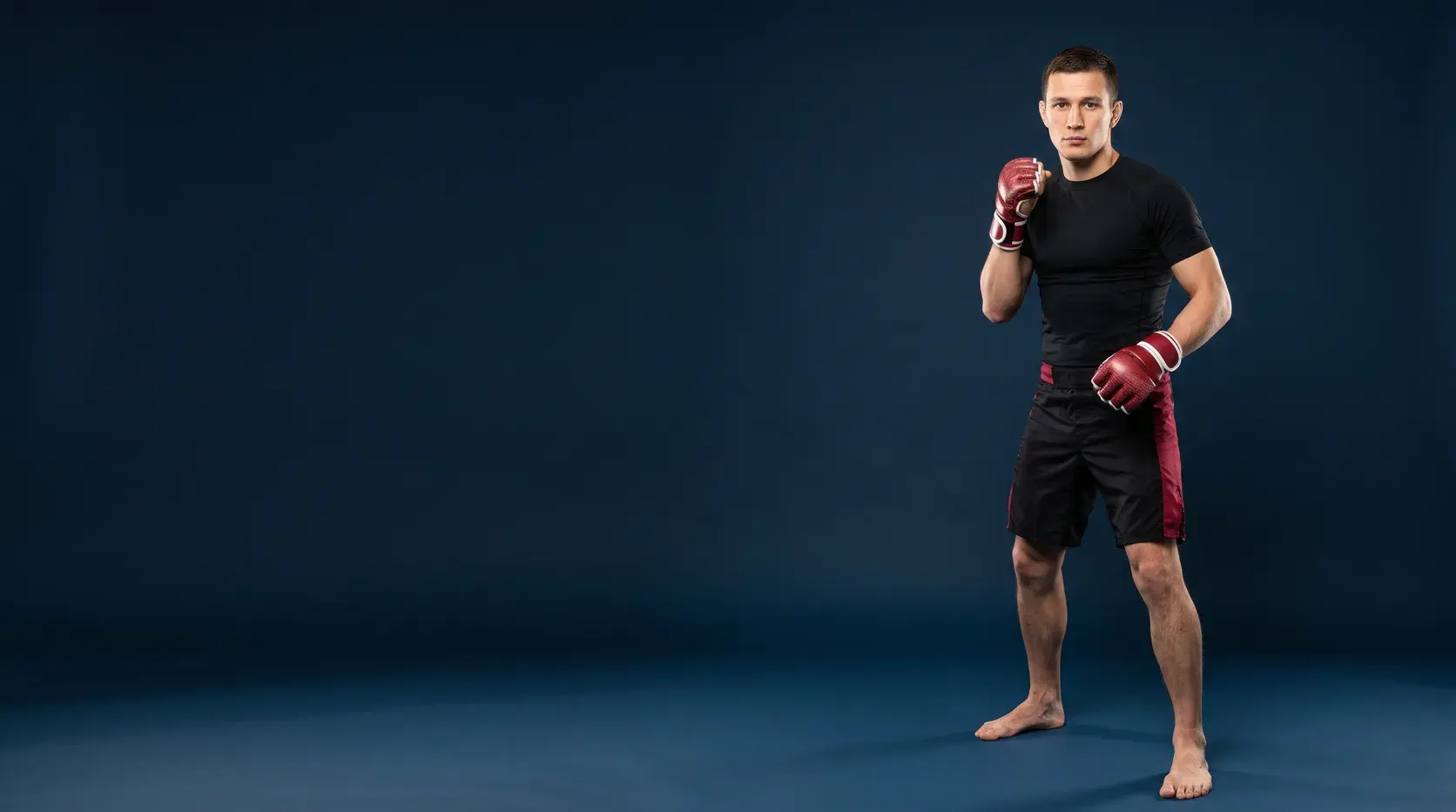 An MMA fighter in a relaxed stance on a midnight navy backdrop, wearing crimson red and white open-palm hybrid gloves, a black rash guard and black fight shorts with crimson side panels, barefoot.