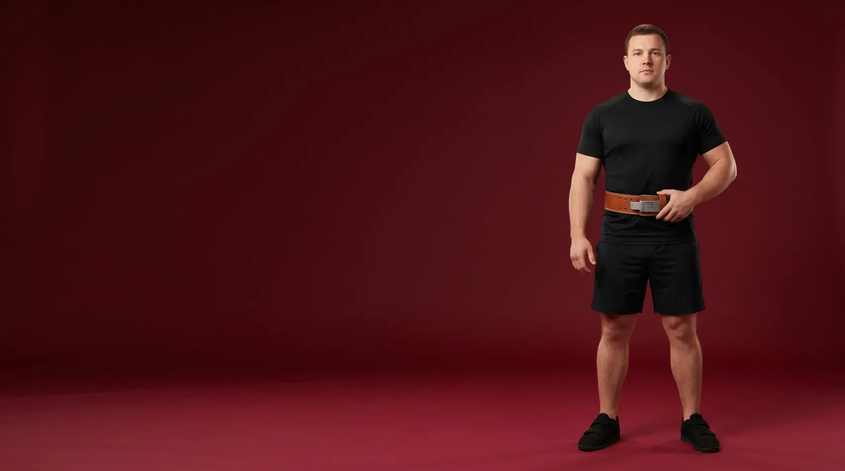 A strength athlete standing on an oxblood backdrop in a tan leather lever lifting belt with a brushed steel lever buckle, one hand resting on the buckle, in a black training top and shorts.